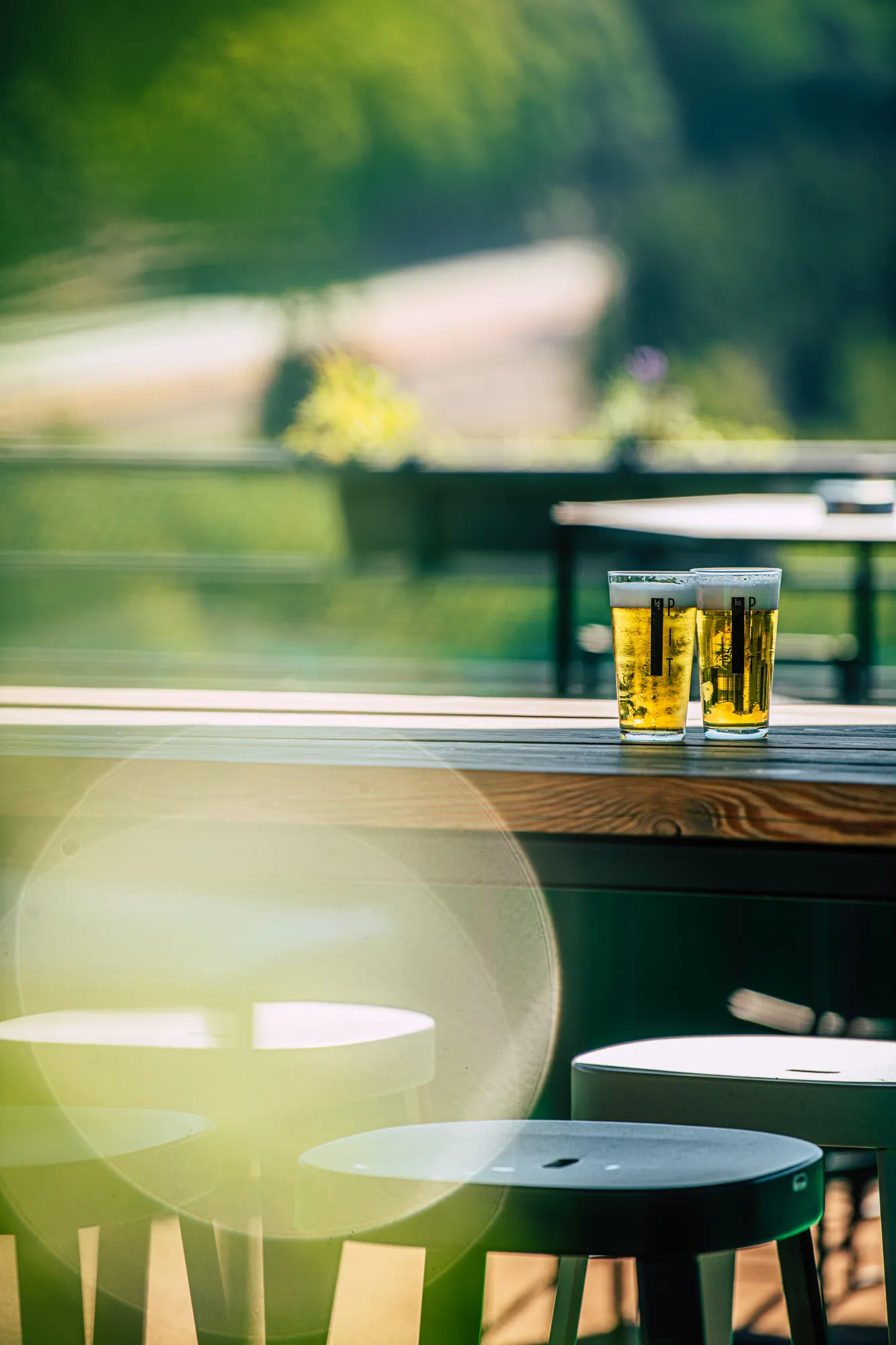 Deux verres de bière sur une table en bois à l'extérieur, avec des chaises floues et de la verdure en arrière-plan.