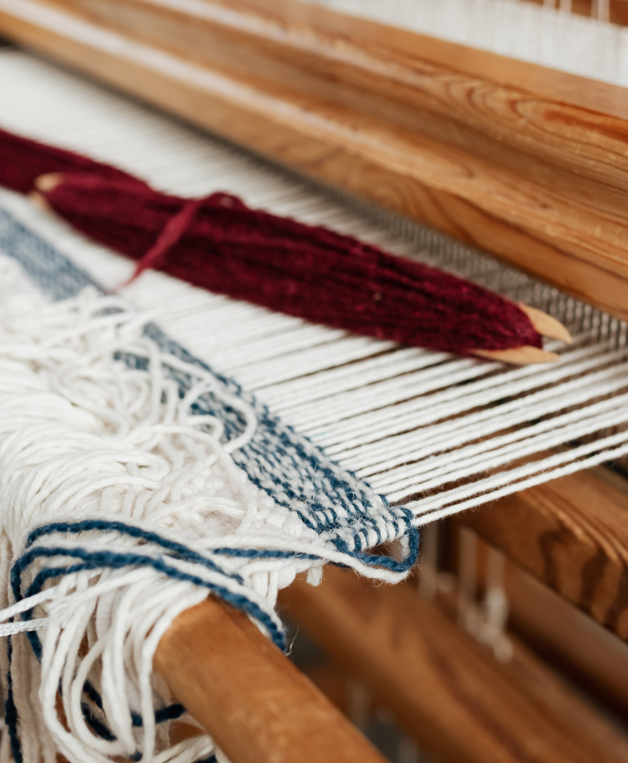 A close up image of a wooden weaving loom and beige, red and blue yarn.