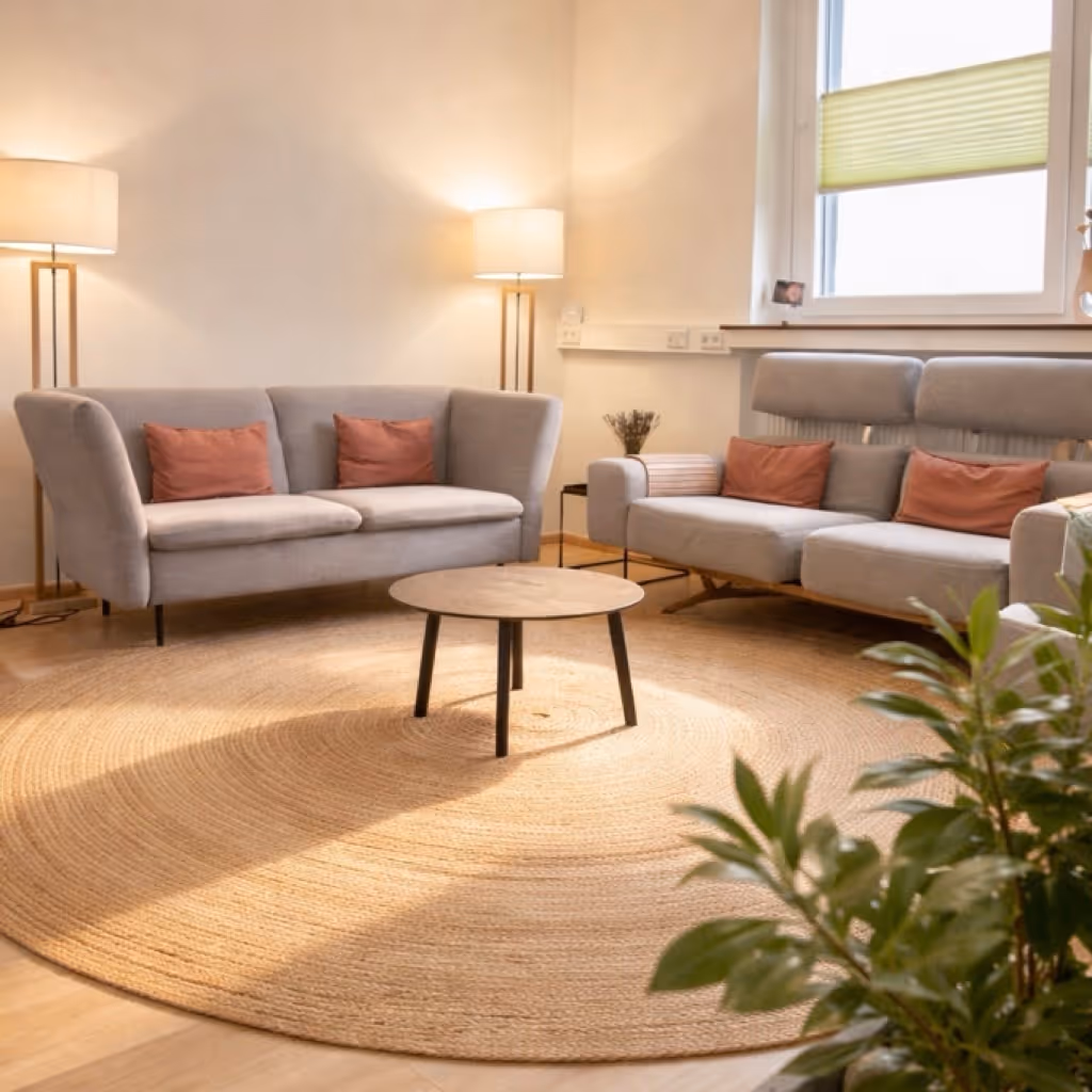 Modern living room with two gray sofas featuring rust-colored cushions, a round wooden coffee table on a large circular woven rug, two floor lamps, and a window with a partially closed blind.