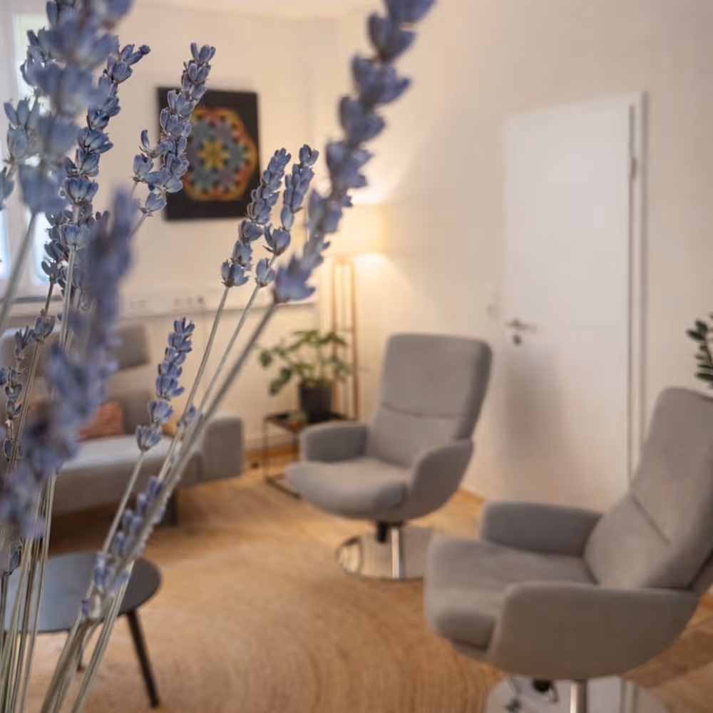 Close-up of lavender flowers with a cozy room featuring two gray armchairs, a sofa, a floor lamp, and a decorative mandala wall hanging in the background.