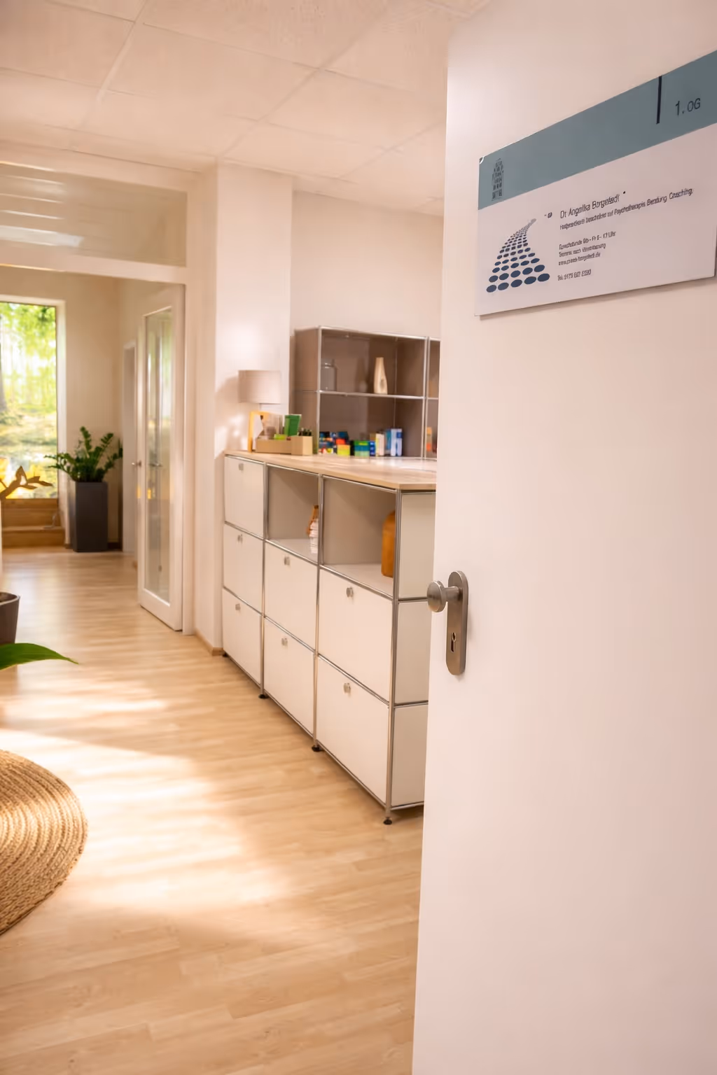 Bright hallway with wooden floor, white storage cabinets, plants, and a door slightly open showing a sign with office information.
