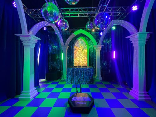 A shiny blue fringed DJ booth sits infront of a stained glass window in this vampire themed mausoleum nightclub. The room is lined with white columns and arches, disco balls hang from the ceiling. The room is bathed in green and purple lights.