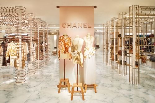 Three Mannequins displayed at the center of a Chanel retail pop-up store. Behind them is a large banner reading Chanel. To both sides are gold cylindrical wire columns integrated with white circular shelving, which hold an assortment of purses, shoes and other accessories.