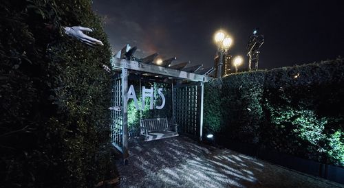 A creepy wooden bench swing with letters mounted above it reading AHS. The swing is surrounded by a hedge maze with hands reaching out from the bushes.