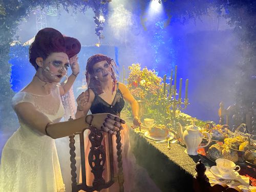 Two women dressed as dolls stand next to a large table. The table is draped in a black cloth and covered in silver trays full of molded food and dying floral arrangements.