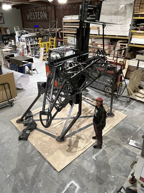 A large metal armature, the internal structure of the bull, standing in the Hotstrike Scene shop. Next to the bull is our kick-ass welder Vimi.