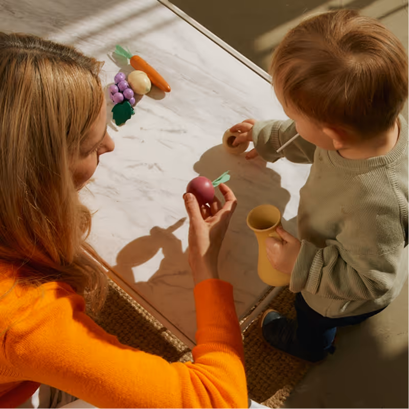 Woman in orange sweater playing with a toddler holding a toy cup and straw at a table with toy vegetables.