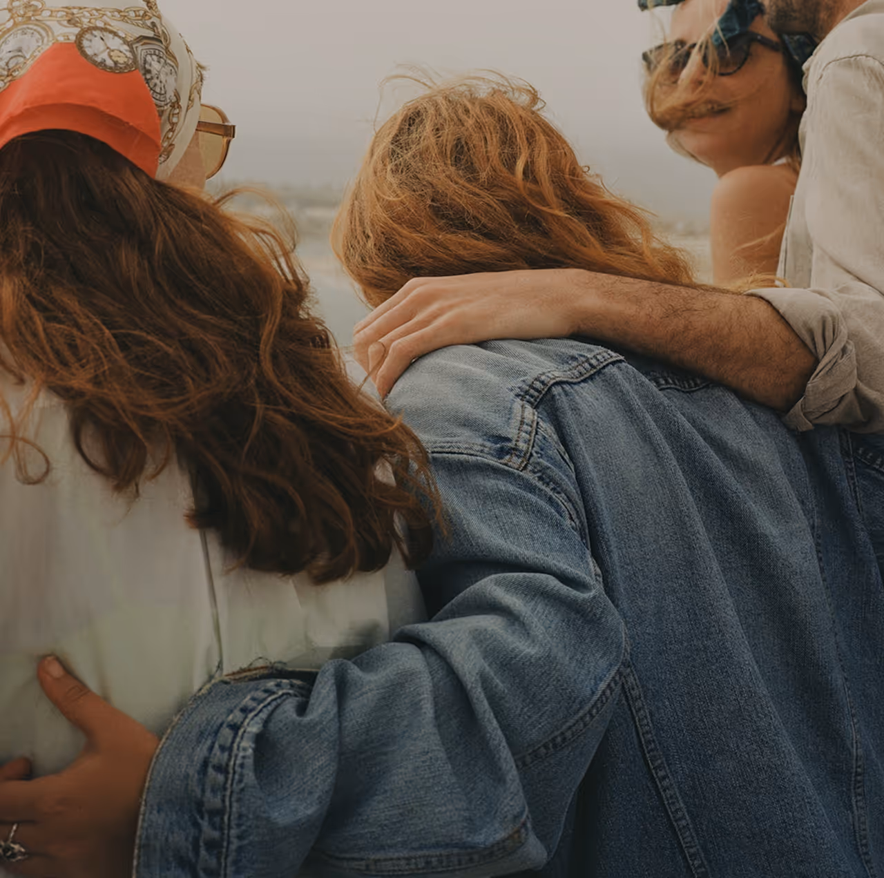 Four friends outdoors with arms around each other, smiling and enjoying a windy day.