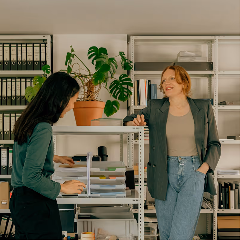 Two women in an office; one organizing papers on a shelf and the other leaning casually and smiling.
