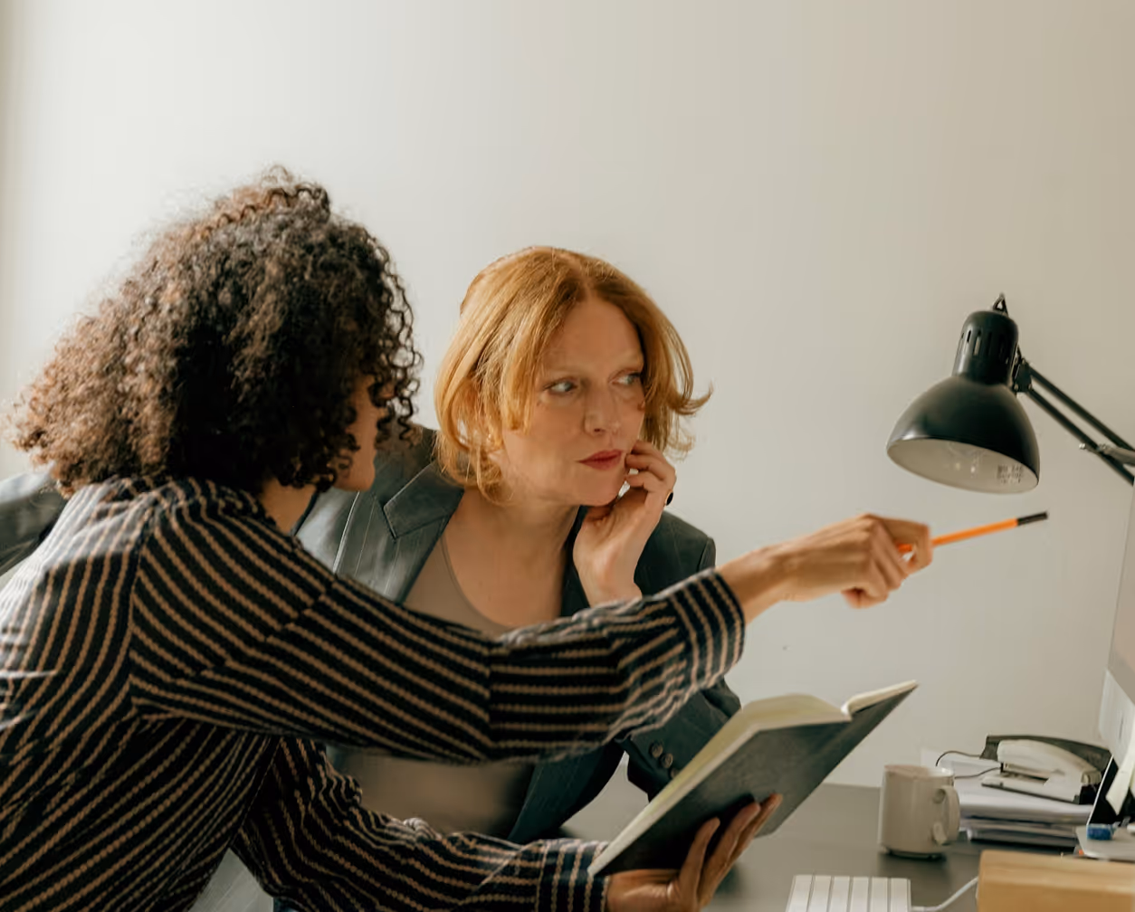 Two women working together at a desk, one pointing at a computer screen with a pencil while the other listens holding a notebook.