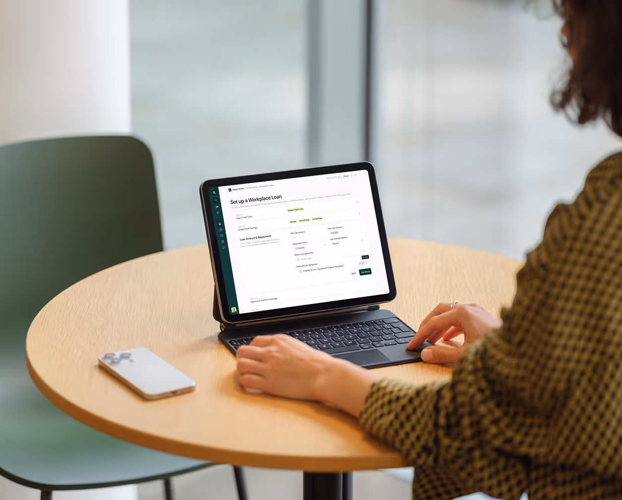 Person working on a tablet with keyboard on a round wooden table, displaying a workplace loan setup form, next to a smartphone.