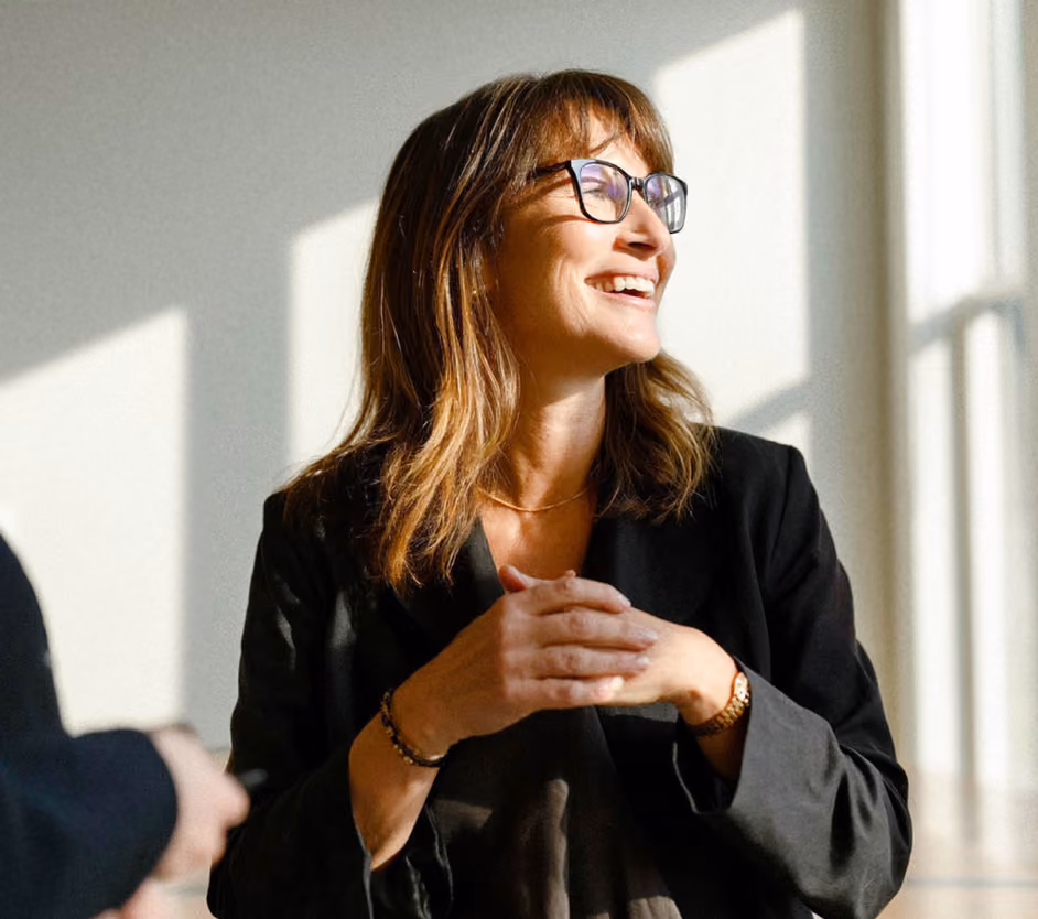 Smiling woman with shoulder-length brown hair and glasses, wearing a black blazer, standing indoors with sunlight casting shadows on the wall behind her.