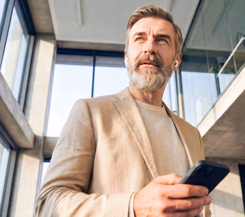 Middle-aged man with gray beard wearing a beige blazer and wireless earbuds holding a smartphone, standing in a modern glass-walled office building.