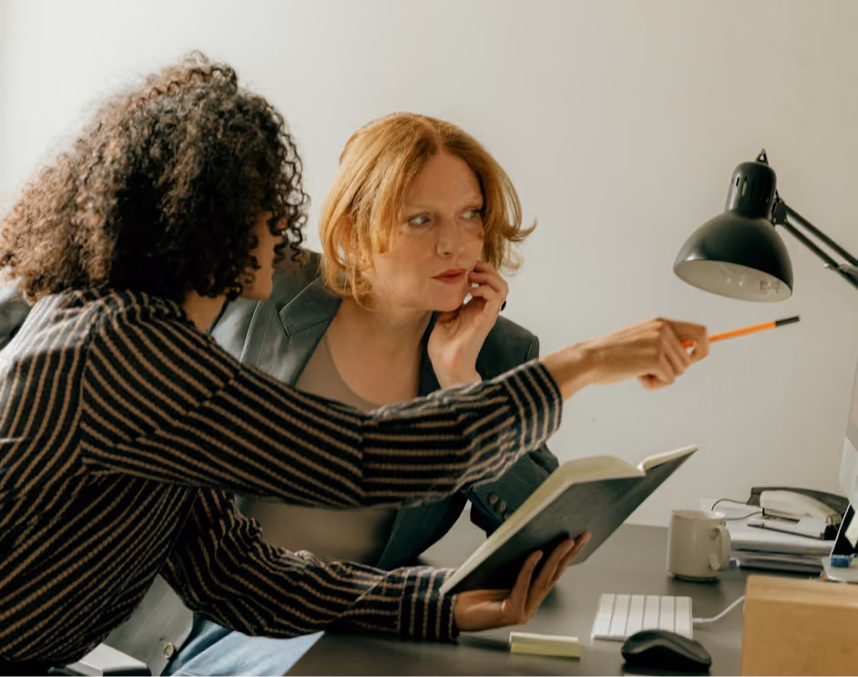 Two women at a desk collaborating; one holds a notebook and points at a computer screen with a pencil while the other listens attentively.