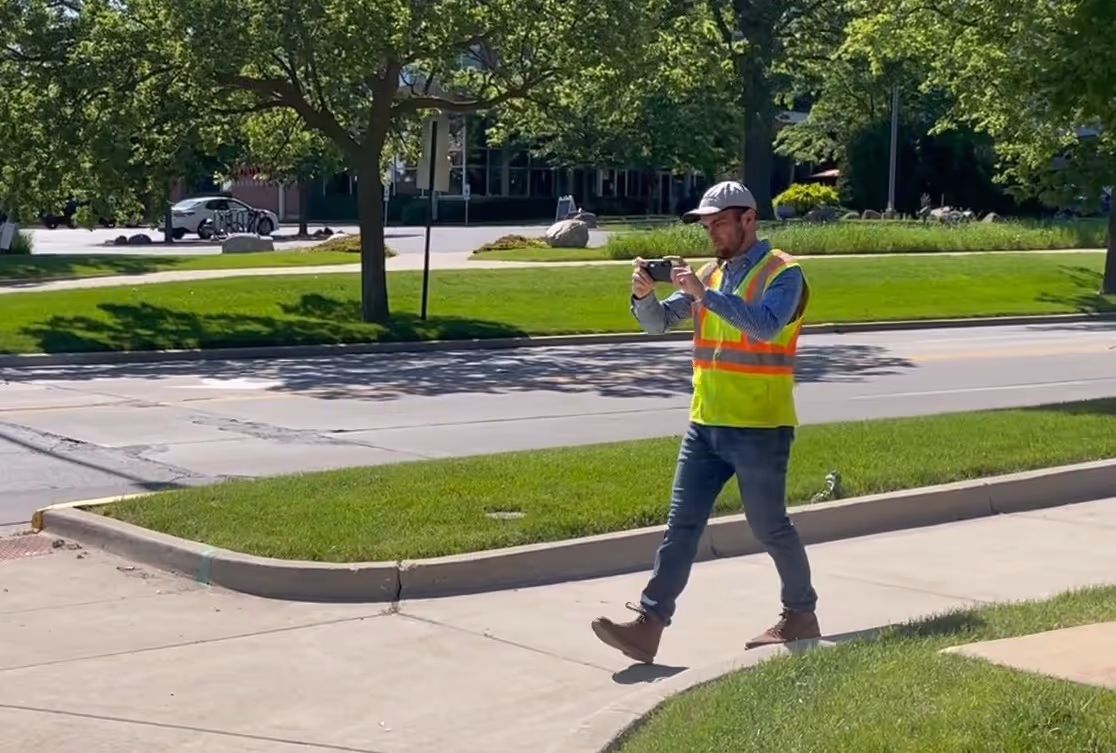 A man in a yellow and orange safety vest walks from right to left on a sidewalk.