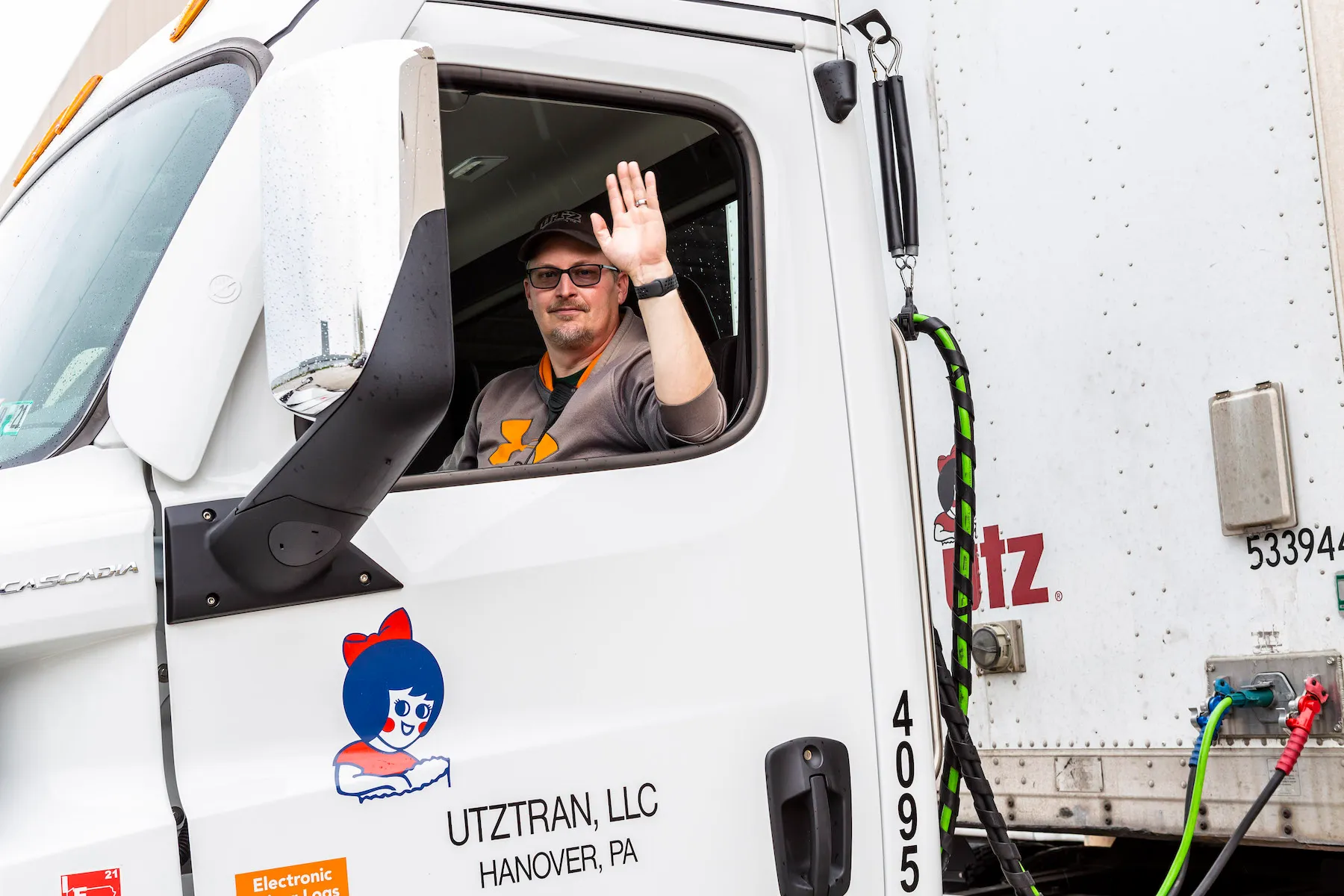 Utz distributor waving at the camera. He's wearing a brown and orange hoodie, sunglasses and brown hat. The truck he is driving has the Little Utz Girl as a decal on the door to the truck.