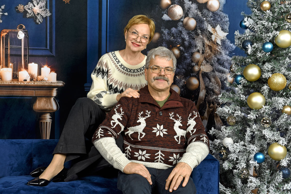 Smiling older couple in festive sweaters sitting on a blue couch beside a decorated Christmas tree with gold and blue ornaments.