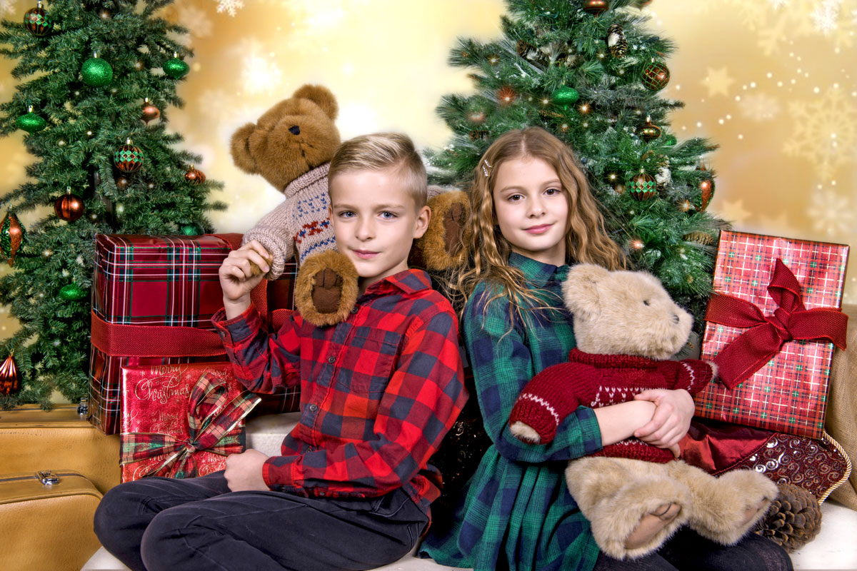 Two children sitting back to back in front of decorated Christmas trees with wrapped presents, each holding a teddy bear.