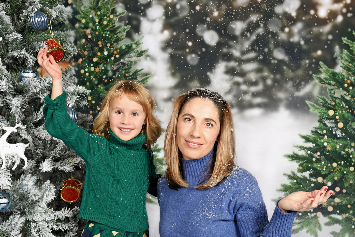 Smiling woman in blue sweater and child in green sweater decorating snowy Christmas tree with ornaments.