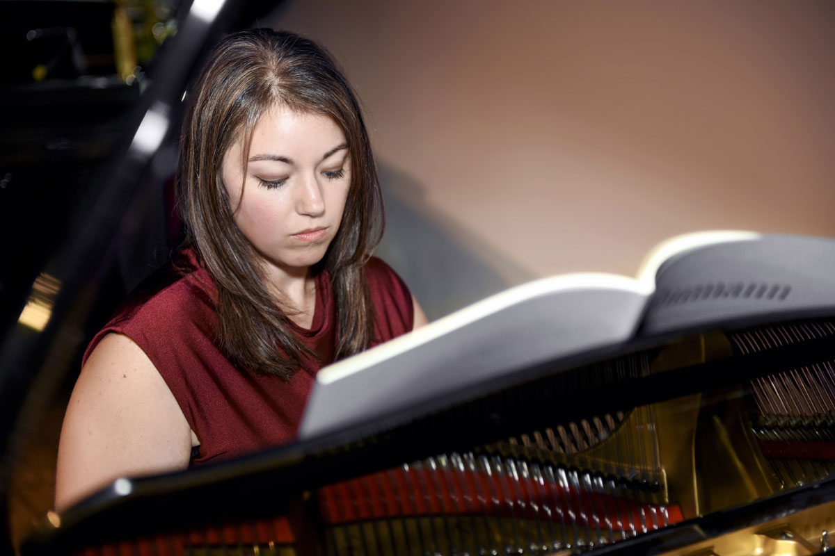 Woman in a sleeveless maroon top focused on sheet music while playing a grand piano.
