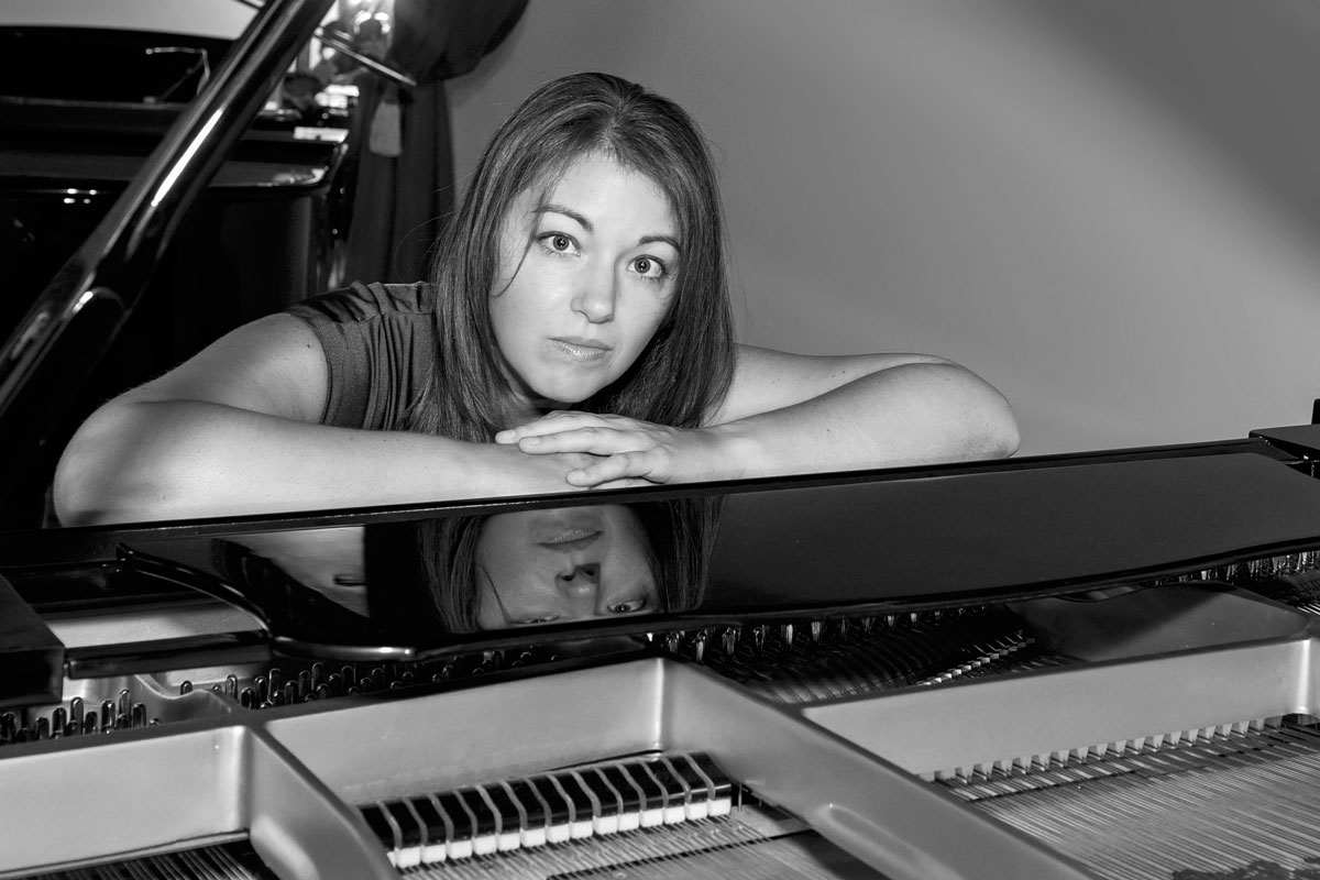 Black and white photo of a woman resting her arms and chin on a grand piano with her reflection visible on the piano's polished surface.