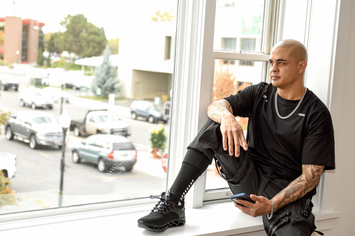 Man with tattoos wearing black clothes sitting on a window sill holding a smartphone and looking outside.