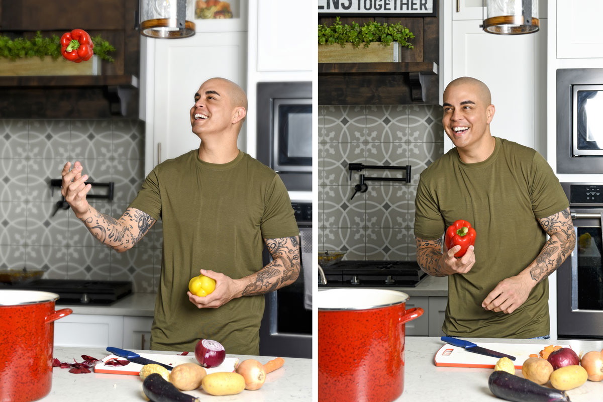 Man with tattoos in an olive green shirt smiling in a kitchen while juggling a yellow and a red bell pepper near a counter with vegetables and a red pot.