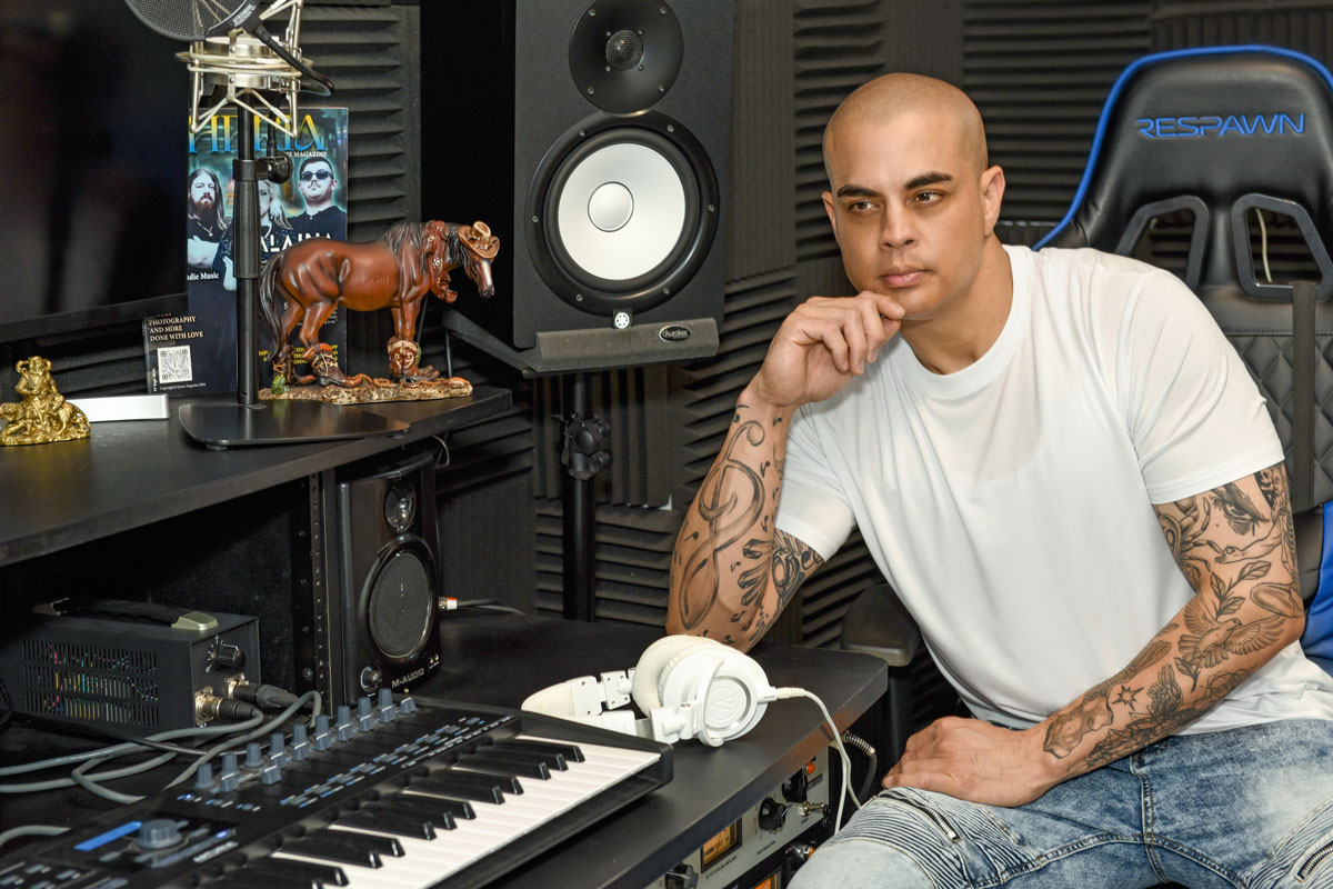 Tattooed man in white t-shirt and jeans sitting thoughtfully in a music studio with keyboard, headphones, and speakers.