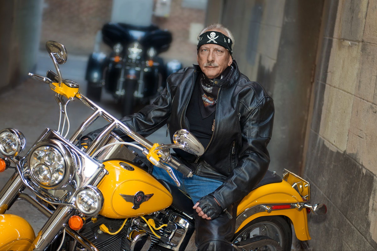 Middle-aged man in a black leather jacket and skull bandana sitting on a yellow motorcycle in an alleyway.