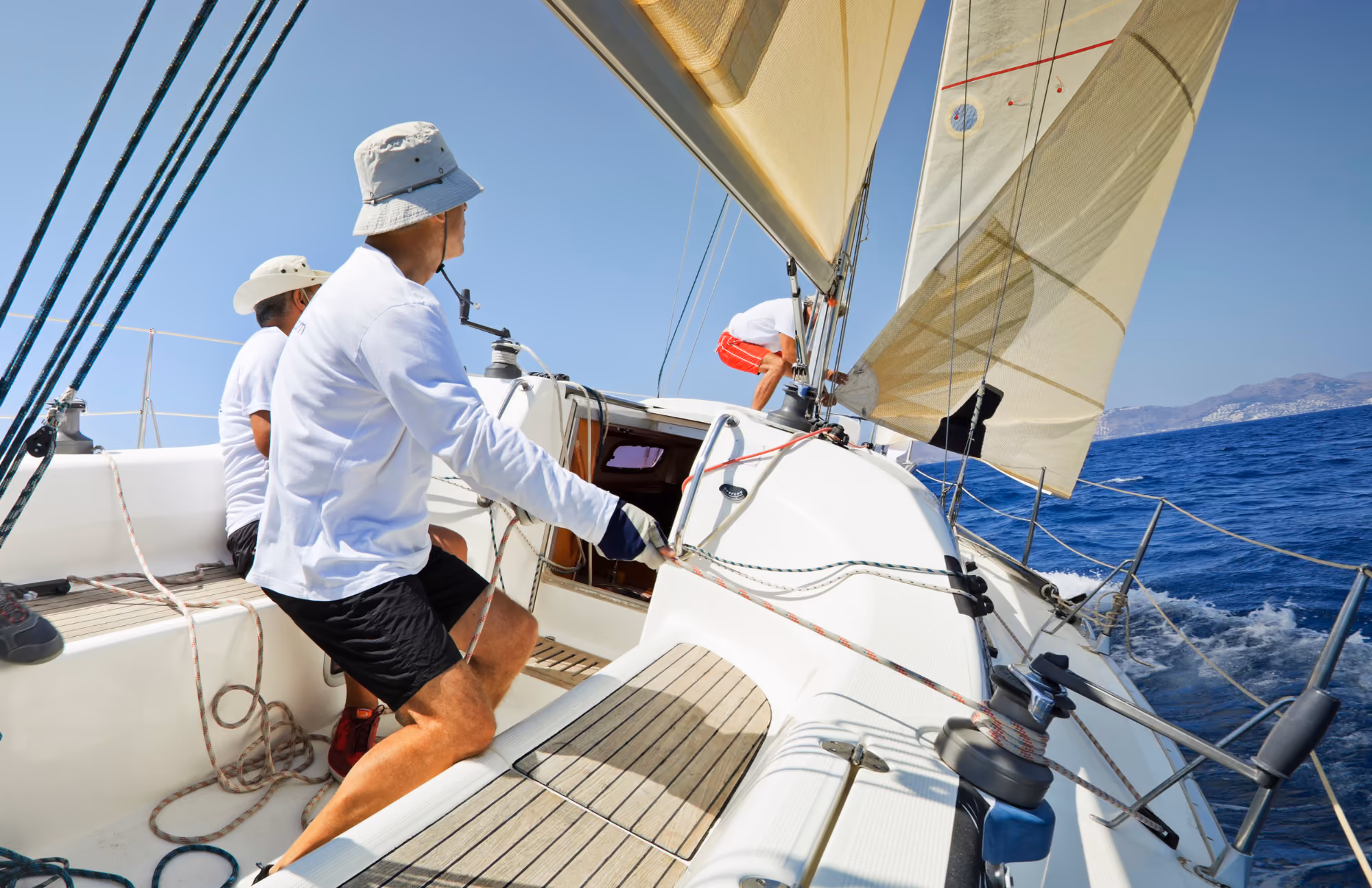 Sailors dressed in white shirts and hats adjusting sails and ropes on a white sailboat sailing on a calm blue sea under clear sky.