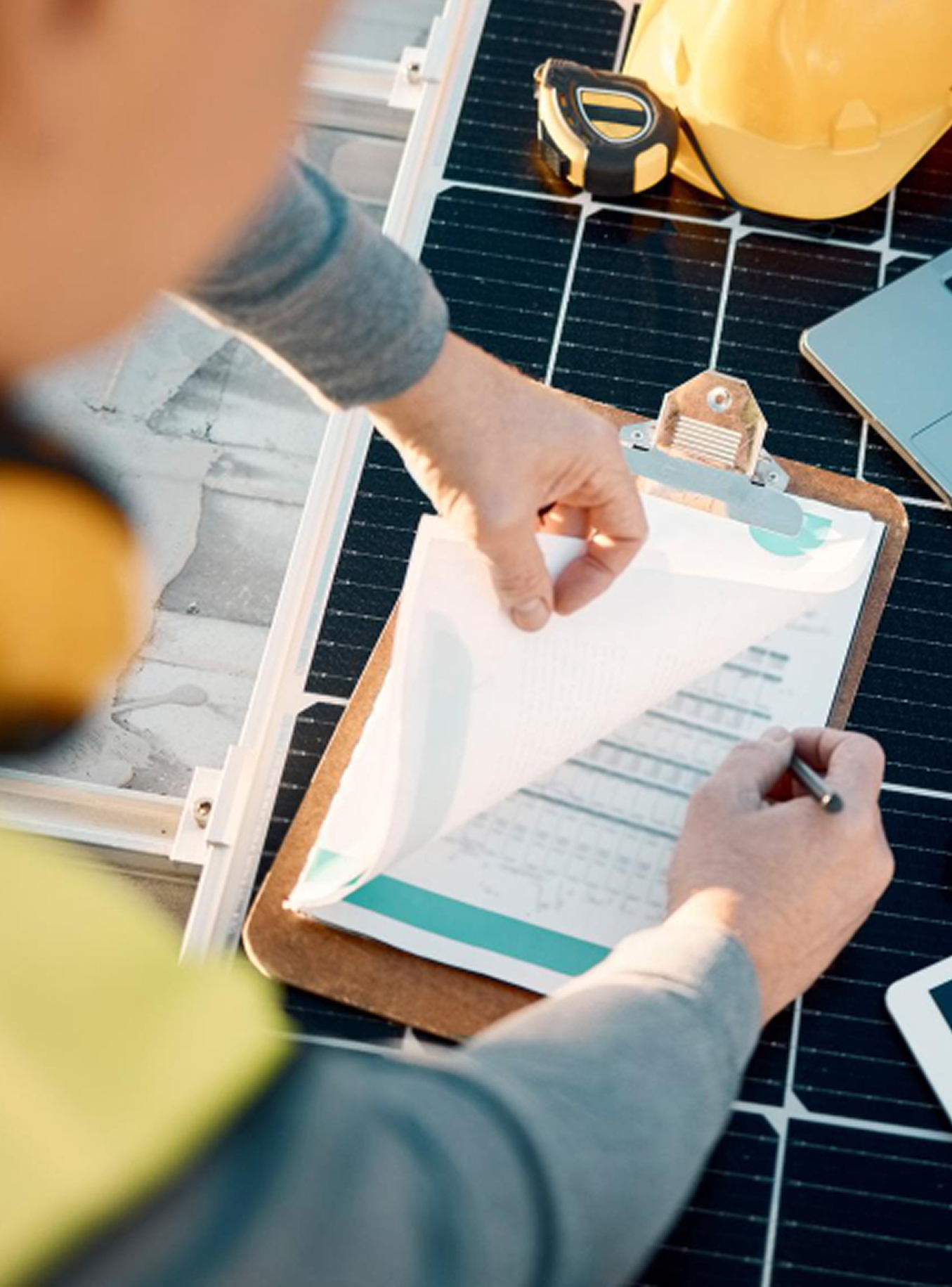 Person wearing safety gear reviewing paperwork on a clipboard placed on a solar panel with a yellow hard hat and measuring tape nearby.