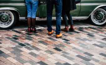 feet and legs of people wearing jeans standing on the side of a green car.