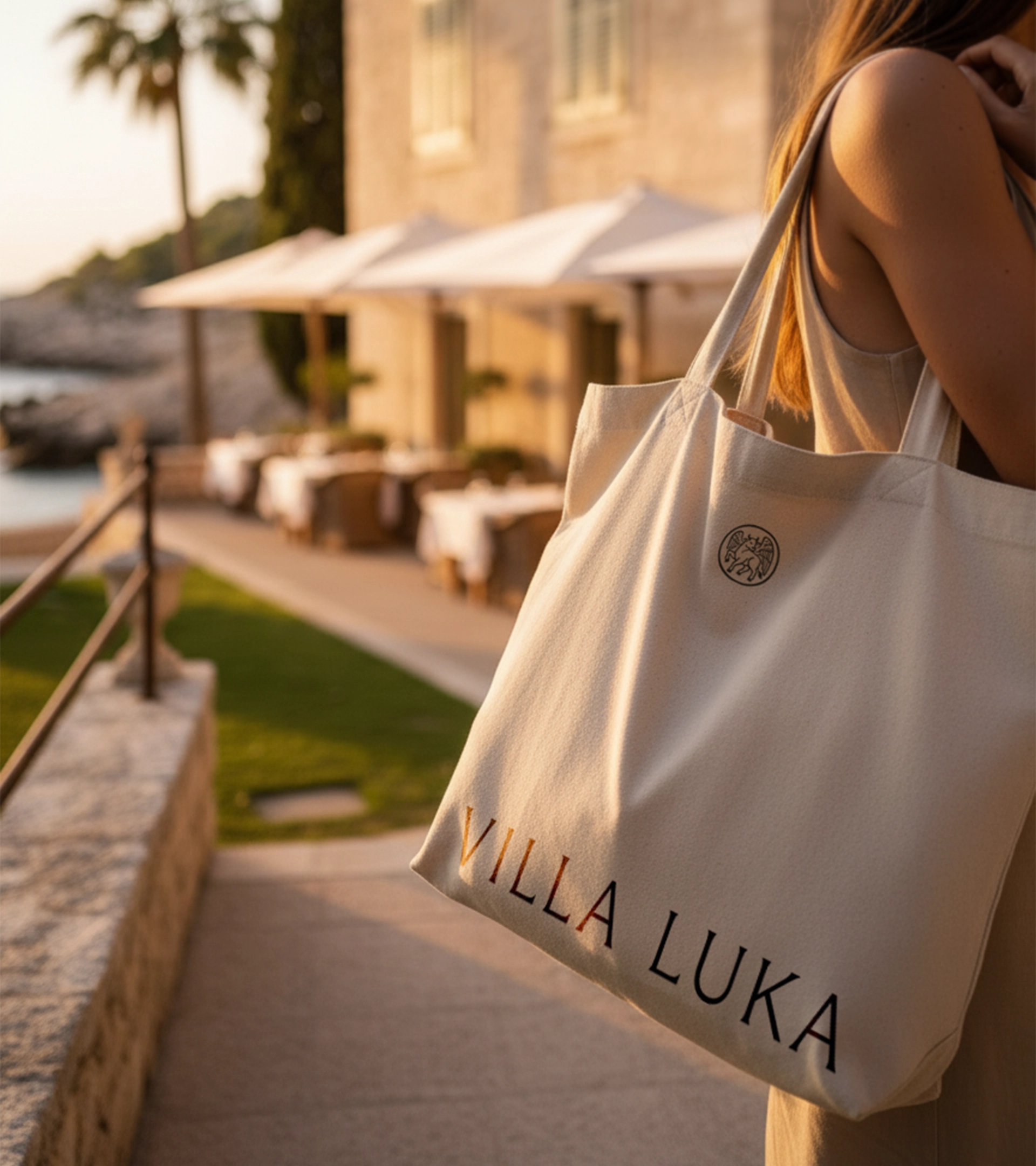 Femme tenant un tote bac beige avec l'inscription « VILLA LUKA » sur une terrasse méditerranéenne ensoleillée donnant sur une piscine et une villa