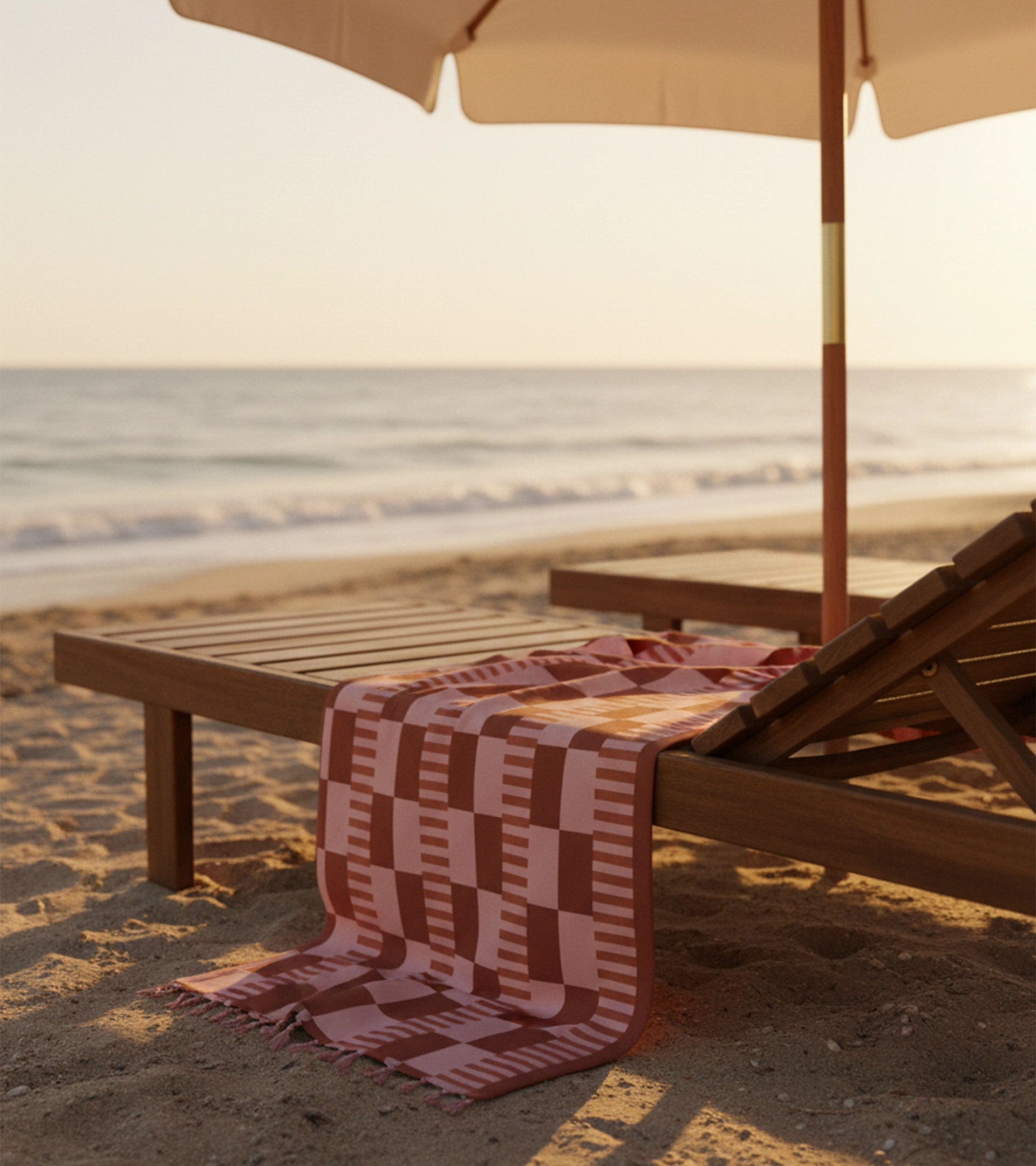Scène de plage au coucher du soleil avec des chaises longues en bois sous un parasol crème, ornées d'un tissu à motifs géométriques dans les tons terracotta et rose drapé sur les meubles