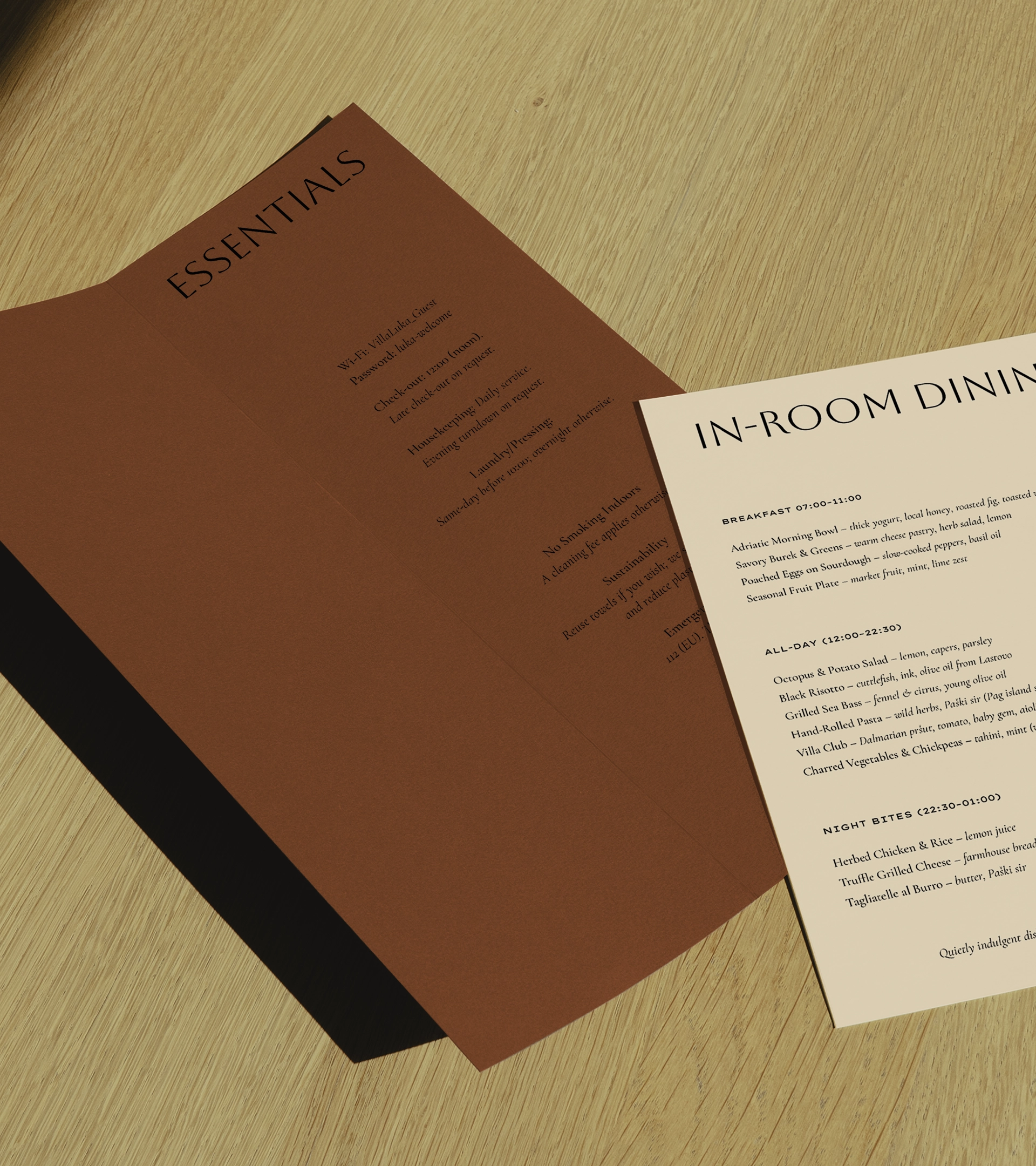 Matériel promotionnel de la Villa Luka, notamment un menu « IN-ROOM DINING » et une brochure rouge « ESSENTIALS » disposés sur une surface en bois clair