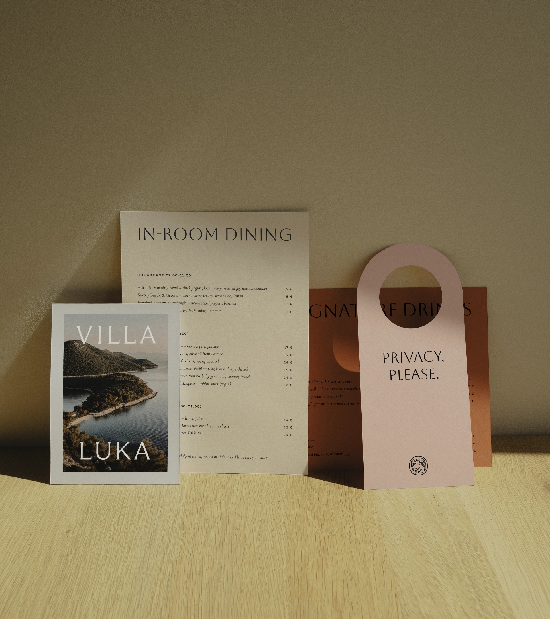 Collection d'articles griffés Villa Luka, comprenant le menu du restaurant, une brochure de photographies côtières et une affichette rose « PRIVACY, PLEASE » avec le logo du taureau ailé