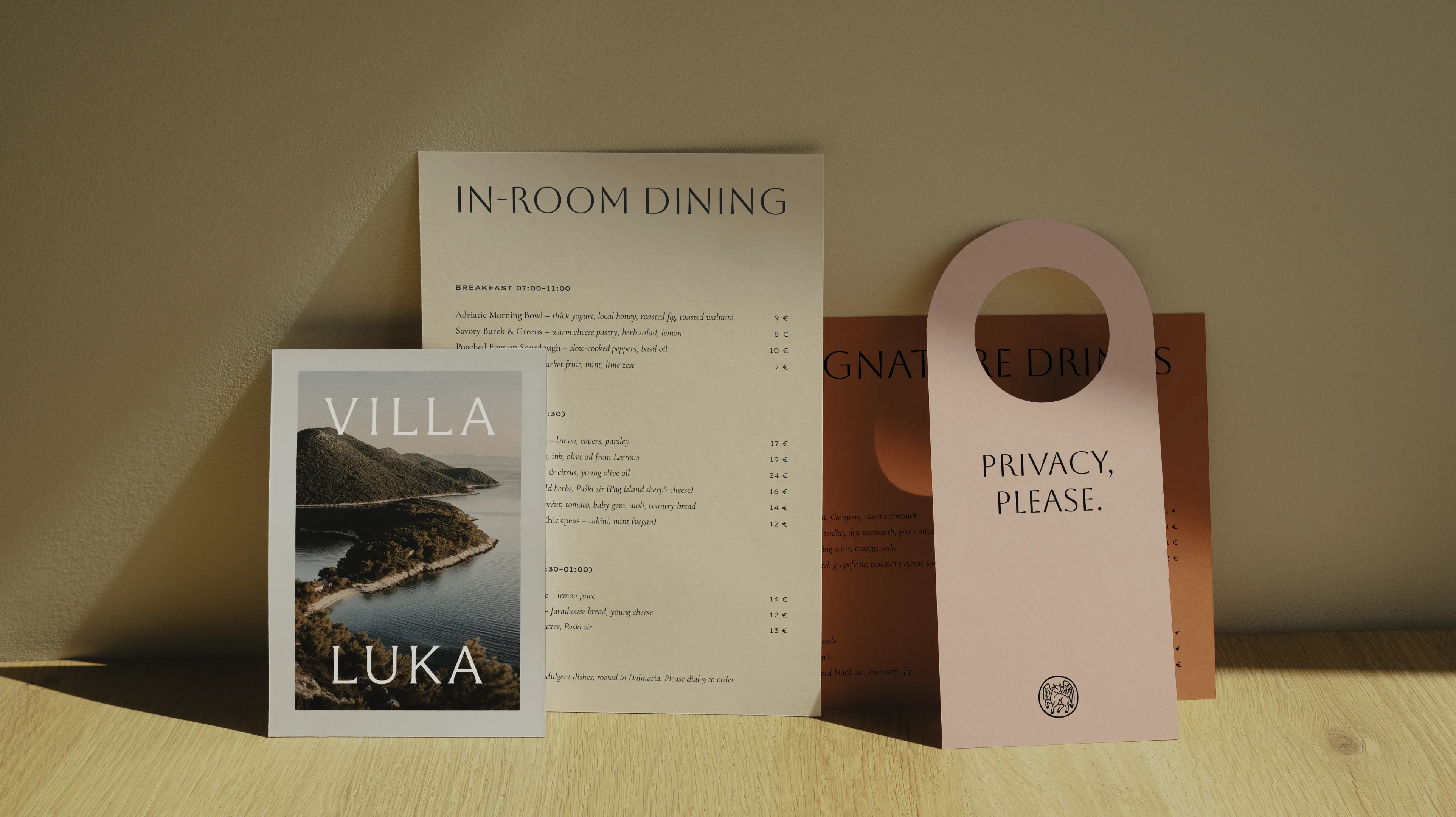 Collection d'articles griffés Villa Luka, comprenant le menu du restaurant, une brochure de photographies côtières et une affichette rose « PRIVACY, PLEASE » avec le logo du taureau ailé