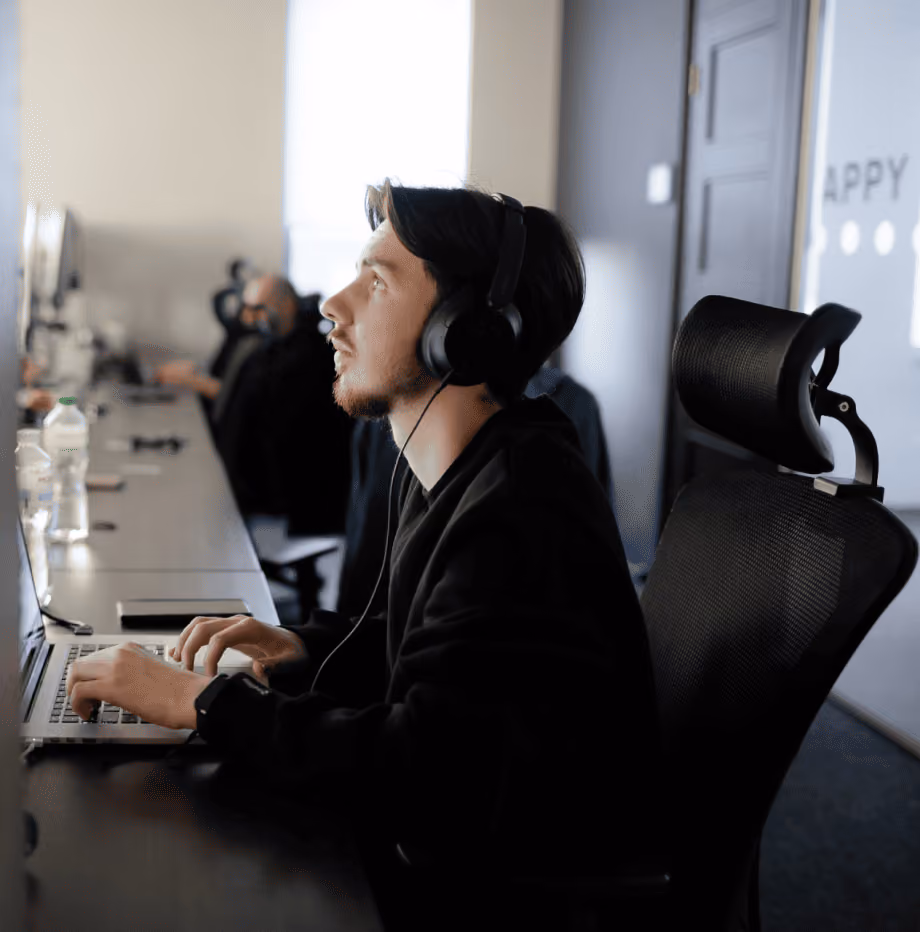 Man wearing headphones working on a laptop in an office with other people in the background.
