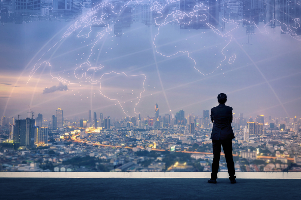 A person in a suit stands on a rooftop, looking out over a sprawling city skyline at dusk, with a translucent, glowing digital map of the world overlaid in the sky above the city.