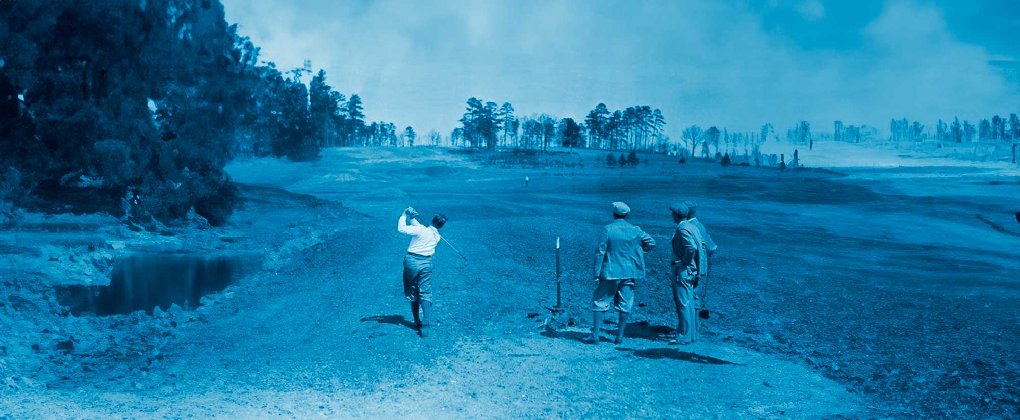 Three men on a golf course with one swinging a club while two watch nearby.