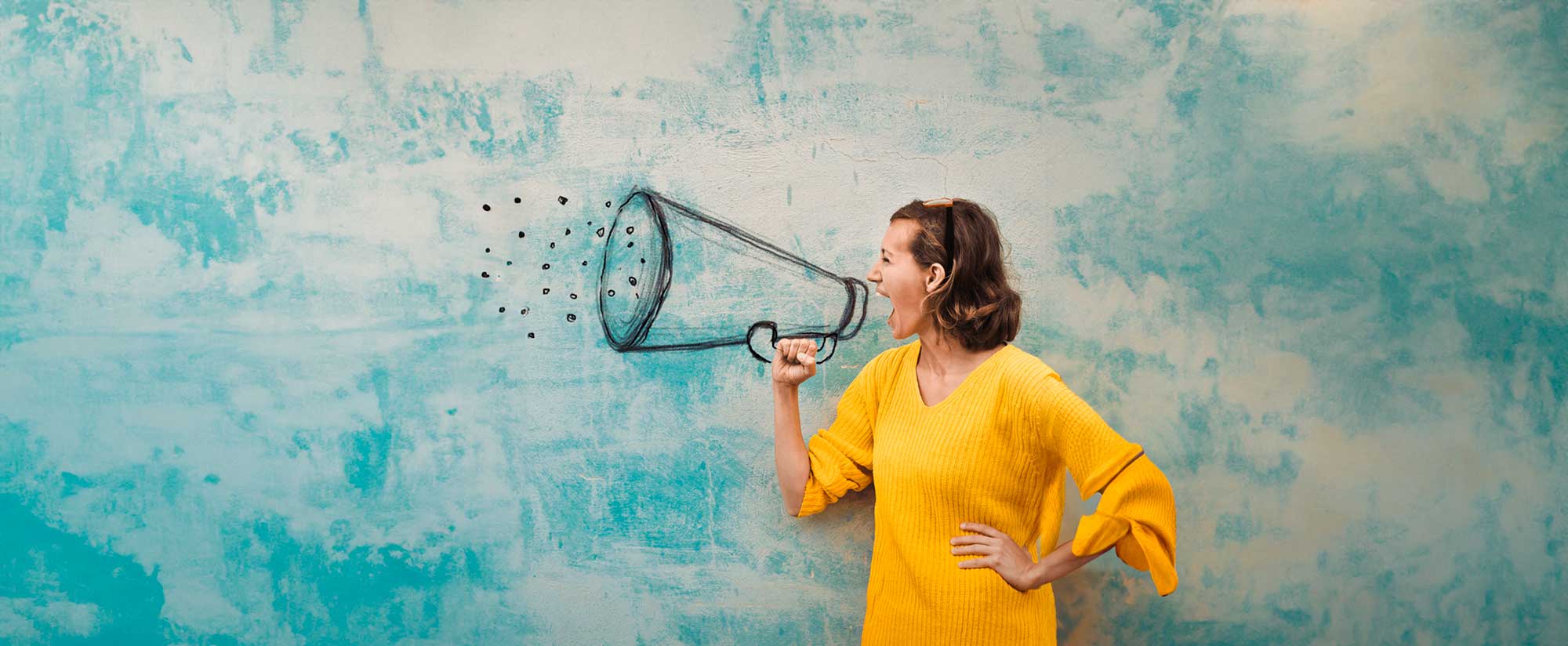 Woman in a yellow sweater pretending to shout into a drawn megaphone on a blue textured wall.