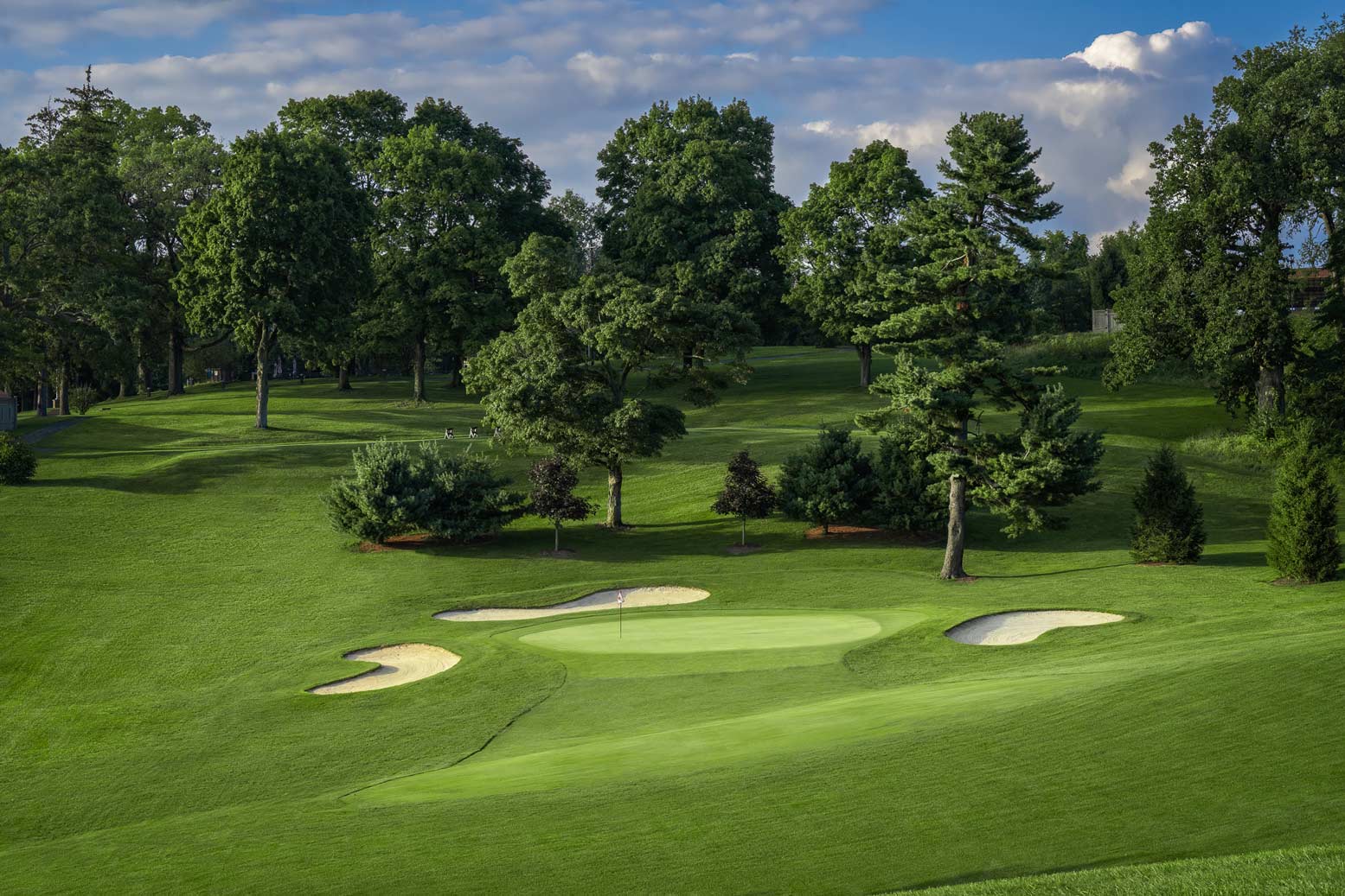 Golf course green surrounded by sand bunkers and lush trees under a partly cloudy sky.