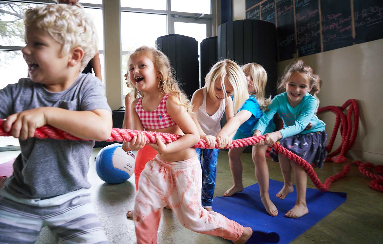 Group of young children playing tug-of-war with a red rope indoors, smiling and having fun.