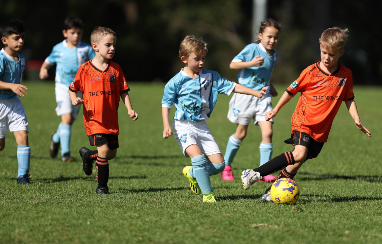 Children playing soccer on grass, with two players in orange jerseys competing for the ball against players in light blue jerseys.