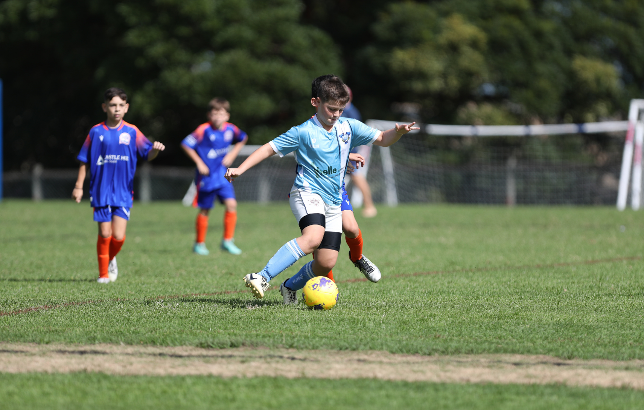 Young boys playing soccer on a grassy field with one boy in light blue jersey controlling a yellow ball while others in blue and orange jerseys watch.