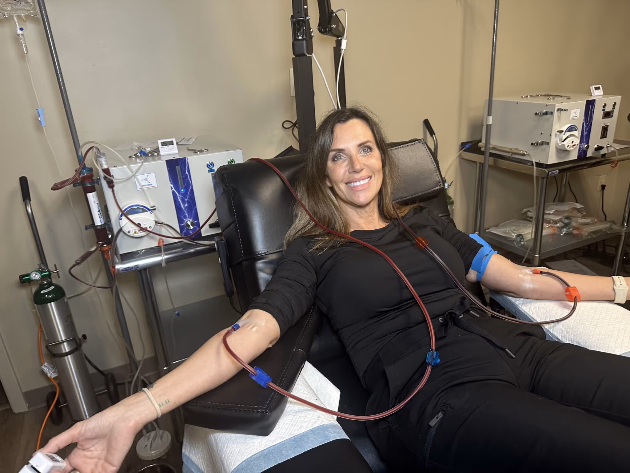 Smiling woman lying on a medical recliner donating blood via tubes connected to both arms.