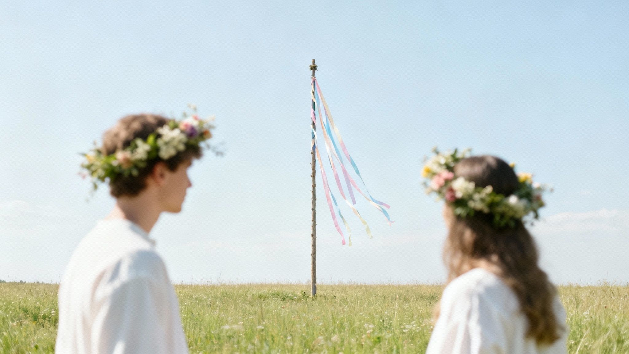 Two people wearing flower crowns look at a colorful maypole in a sunny, green field.