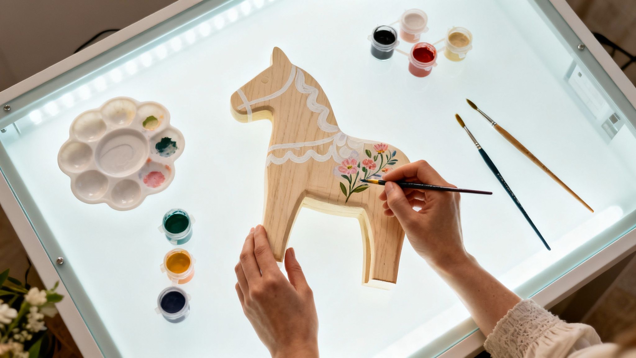 A person painting a wooden horse with delicate floral designs on a light table, surrounded by art supplies.