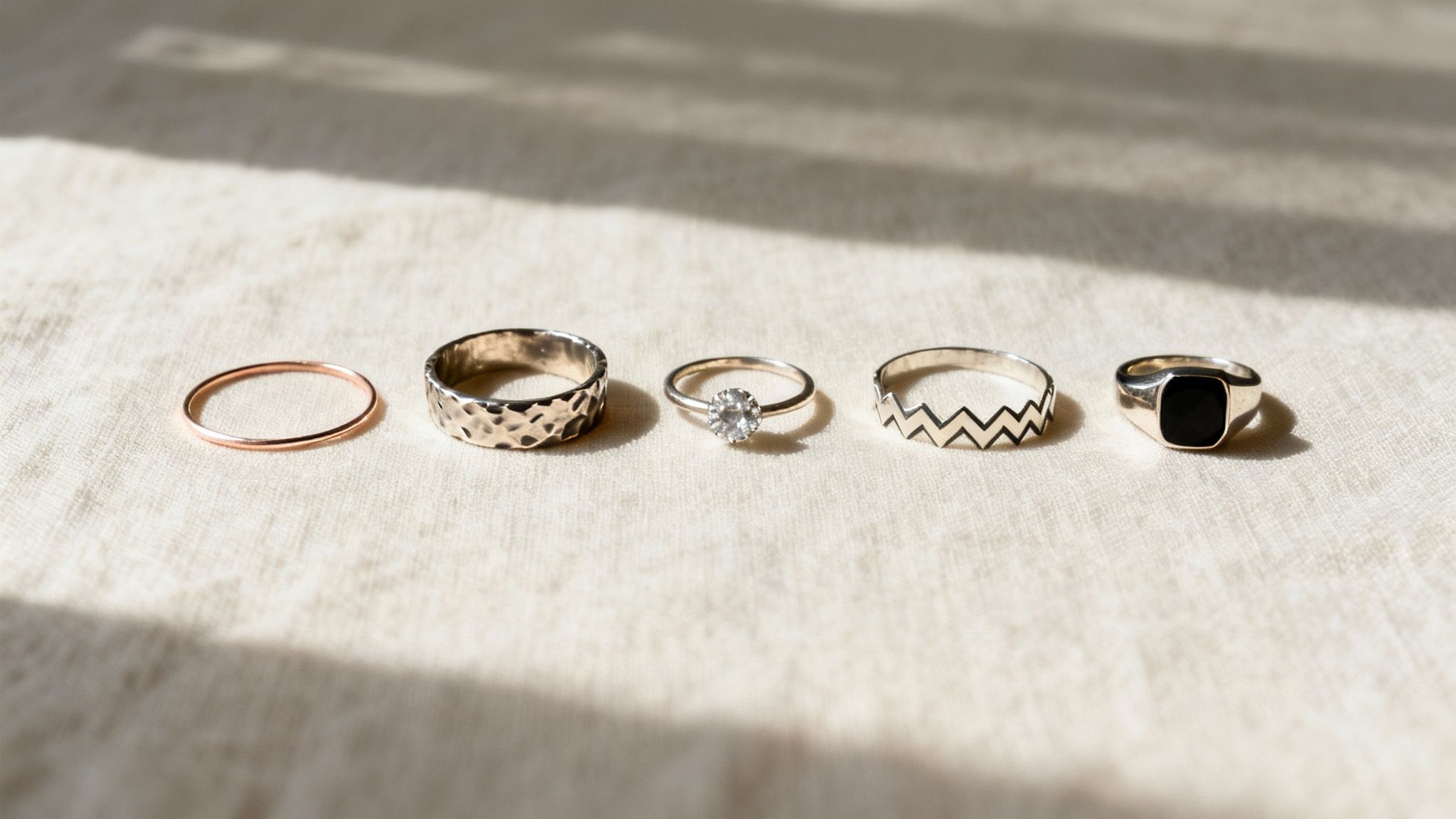 Five diverse rings, including a rose gold band, hammered silver, solitaire diamond, zig-zag, and black signet, on fabric.