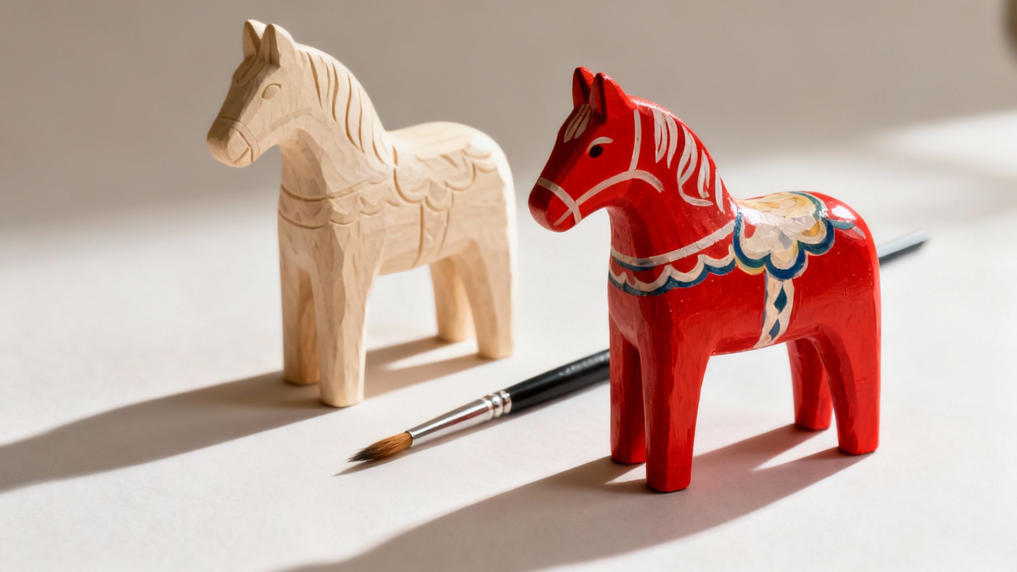 Two Dala horses, one unpainted and one red with patterns, with a paintbrush.